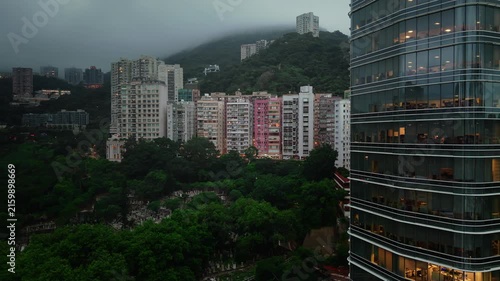 Aerial drone view of a dense residential area in Hong Kong, featuring high-rise apartment buildings, local sports facilities, roads, and lush green hills under cloudy weather.