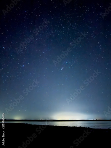 Obraz na plátne A breathtaking night sky filled with countless stars, including the Orion constellation, glowing above a dark silhouette of a lake and horizon with distant city light pollution