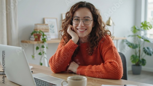 Smiling young woman with curly hair sitting at desk working on a laptop computer setup with wearing glasses orange sweater sitting wooden cheerful woman