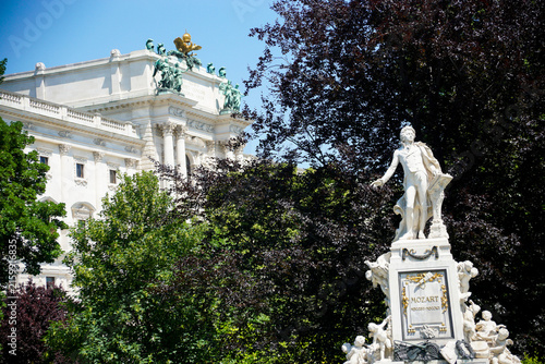 White Mozart monument surrounded by lush trees and formal greenery, with a grand classical building in the background, ideal for travel, culture and heritage content.
