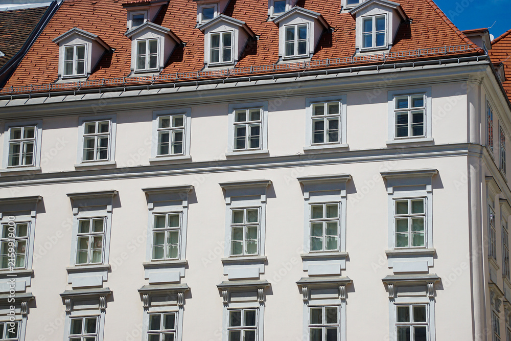 Obraz premium Low-angle view of a historic white building with repeating windows, decorative facade details, dormer windows, and a red tile roof against a clear blue sky.