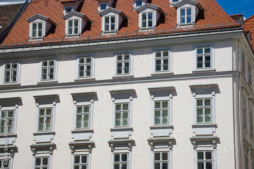 Low-angle view of a historic white building with repeating windows, decorative facade details, dormer windows, and a red tile roof against a clear blue sky.