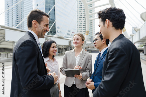 Group of diverse business professionals smiling and discussing plans outdoors in a modern city setting, standing under a contemporary structure with glass buildings in the background.