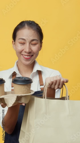 Vertical Screen : Happy smile Asia waitress wear apron friendly looking at camera giving coffee or tea delicious food in craft paper bag to customer isolated on yellow background. Business takeaway.
