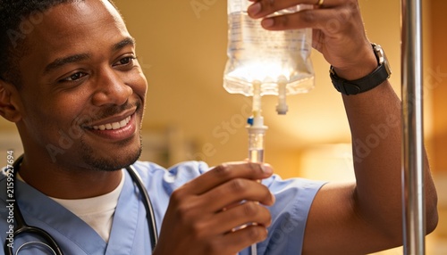 Adjusting healthcare worker in blue scrubs attaching IV tubing at patient bedside, holding IV bag