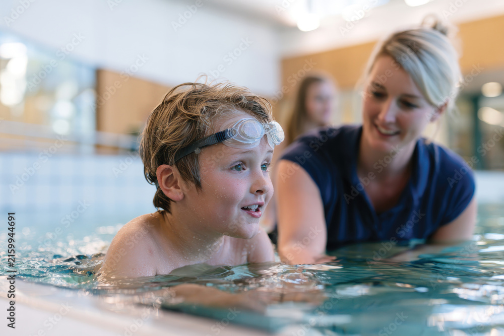 Fototapeta premium A happy child wearing swim goggles engages in swimming lessons, showcasing enthusiasm for learning in a vibrant aquatic environment under the attentive guidance of a friendly instructor.
