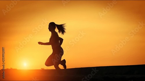 Silhouette of an athletic woman running along a horizon line against a vibrant orange sunset sky.