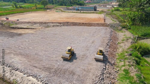 Aerial view of toll road construction with yellow road rollers compacting gravel on a site surrounded by green fields in Muntilan, Central Java, Indonesia.