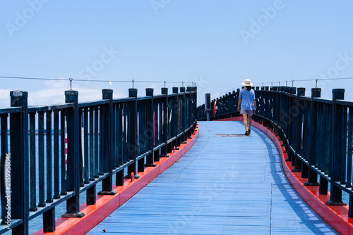 Woman with Sun Hat Walking on Bright Blue Wooden Coastal Footbridge