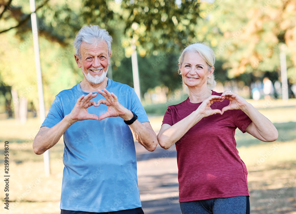 Fototapeta premium Smiling active mature middle aged couple people posing together forming heart shape with hands in the park