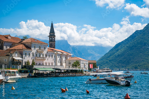 Waterfront view of Perast, Montenegro, showing a stone church bell tower, terracotta rooftops, moored boats on the blue bay, and mountains beneath clouds