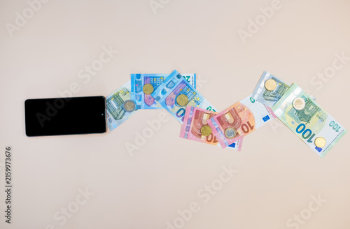 Black smartphone showing an online shopping or payment interface beside scattered euro banknotes and coins on a beige surface, depicting digital purchases