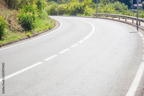Winding paved road curving through lush green roadside vegetation, with white lane markings, metal guardrail, railing, and distant houses