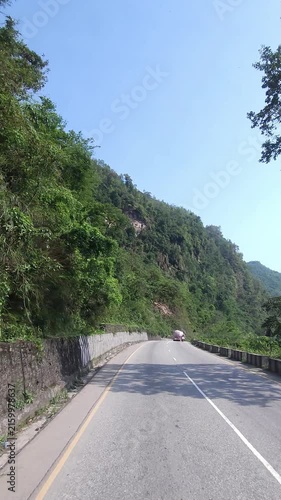 Vertical travel footage showing a winding mountain road passing through dense green forested hills in Nepal. Narrow roadway beside a concrete roadside barrier.