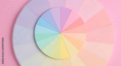 A circular color wheel composed of various pastel paper segments, arranged in a gradient pattern on a soft pink background, top-down view