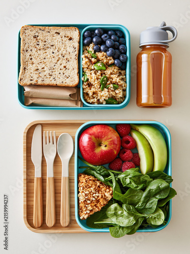 School lunch box flat lay packed lunch reusable container sandwich fruit utensil spinach bottle grain meal prepared for student break on beige surface