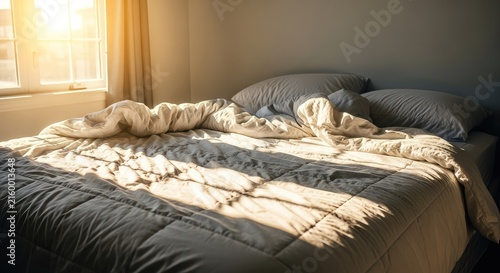 A cozy unmade bed with a fluffy white comforter is bathed in warm morning sunlight from a window, creating a serene and restful atmosphere, morning, textile, sheets