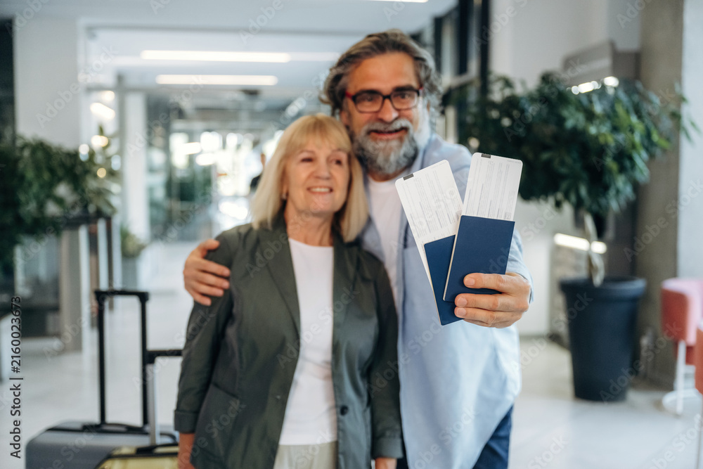 Fototapeta premium Senior couple smiling and holding travel tickets in a modern airport terminal with luggage and greenery in the background, ready for their journey