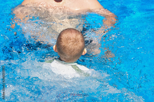 Young boy in a white rash guard practices swimming toward an adult with outstretched arms, illustrating swim lessons, water safety, swimming skills, and learning to swim.