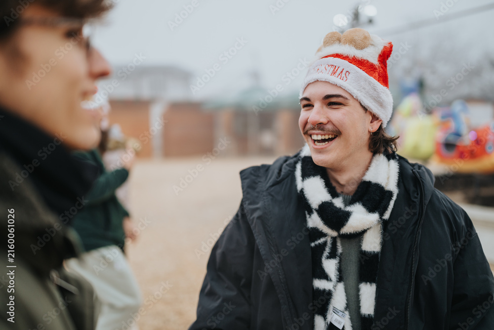 Fototapeta premium A young man wearing a Santa hat smiles and laughs while talking with friends outdoors. The festive scene shows casual winter clothing and a cheerful atmosphere near an amusement ride.
