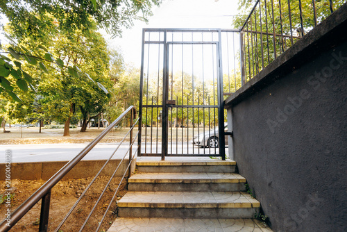 Outdoor metal gate at top of tiled steps with handrail beside sunlit park, framed by concrete wall, vertical bars, and urban entrance details in warm afternoon light