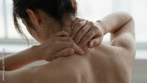 Young woman holding her neck in pain and discomfort at home, grimacing from neck ache and tension while seeking relief and self-care in a bright modern living space