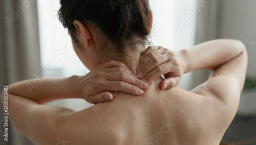 Young woman holding her neck in pain and discomfort at home, grimacing from neck ache and tension while seeking relief and self-care in a bright modern living space