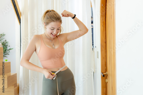 Obraz na plátně Happy young woman measuring waist with tape measure flexing arm muscle showing w