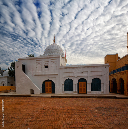 View of the white Gurudwara Patti Sahib with its distinctive dome and arched doorways set against a dramatic cloudy sky in the brick courtyard of Nankana Sahib, Punjab, Pakistan.