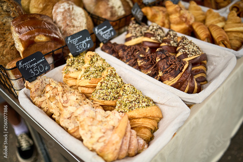 Variety of fresh baked pastries including croissants, cinnamon rolls, and pistachio-topped sweets displayed on trays with price tags at bakery or farmers market stall.