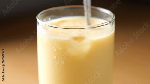 Close-up shot of clear liquid being poured into a tall glass filled with iced creamy beverage, showing condensation and a refreshing summer drink concept.