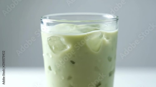 Close up slow motion shot of water being poured into a tall glass of iced creamy green beverage, likely a matcha or green tea latte, showing refreshing condensation drops.