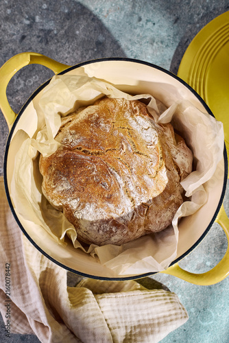 Top view of a golden crusty artisan loaf resting in a yellow enameled cast iron pot on parchment paper. Traditional homemade baking concept.