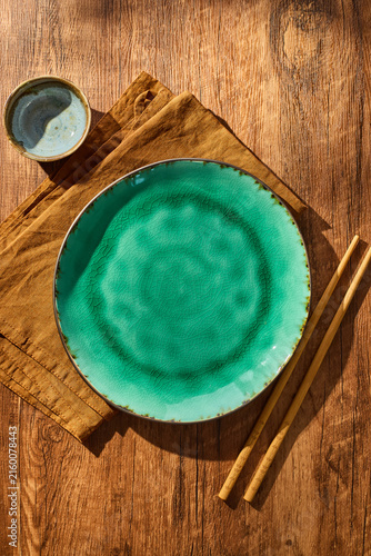 Empty turquoise ceramic plate with brown napkin, wooden chopsticks and small bowl of soy sauce on rustic wooden table. Asian dining setup, food mockup, overhead view with natural lighting.