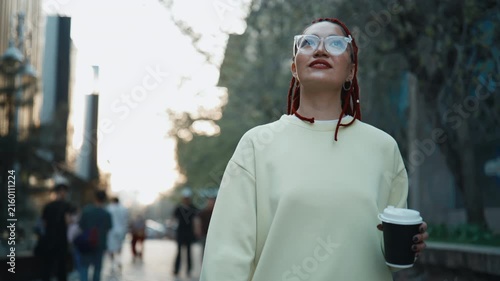 Woman with red braids and glasses walking through city street holding coffee cup smiling