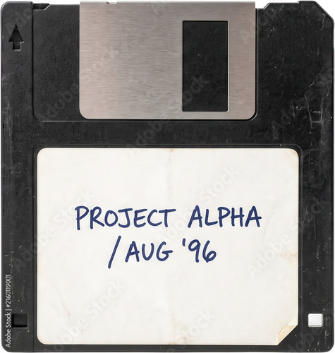 An old black floppy disk with a metallic shutter and a white paper label handwritten with blue ink text displaying project alpha and a date stamp from august nineteen ninety six