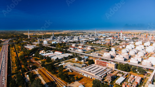 Oil refinery tank farm beside highway under dark clouds. Drone view shows white tanks, process towers, pipelines, rail lines, warehouses and smoke near open fields.