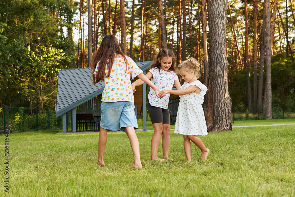 Fototapeta premium Three children dance and hold hands while standing on green grass in a park. The sun is low in the sky, creating a warm atmosphere. Trees surround them