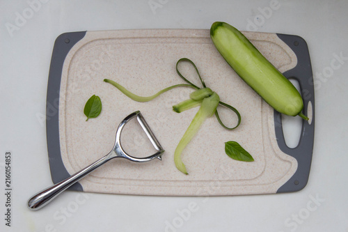Fresh peeled cucumber, cucumber peels in the form of a bow pattern and peeler on cutting board. Concept of preparing a vegetable meal, decoration of dishes. Top view.