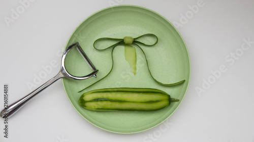 Fresh peeled cucumber, cucumber peels and peeler knife on a green flat plate. Concept of preparing a vegetable meal, decoration of dishes. Cleaning and cutting vegetables. Top view.