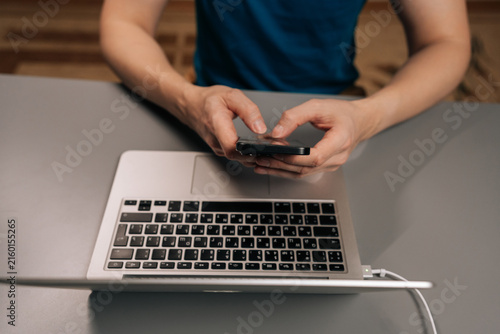 Top view of male hands typing on smartphone while sitting at desk with open laptop, showing busy professional managing messages, tasks and online communication in dark modern home office.