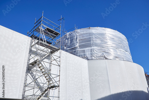 Building renovation construction site with scaffolding and protective sheeting around a large structure. Exterior construction work features temporary scaffolding, access stairs, weatherproof covering