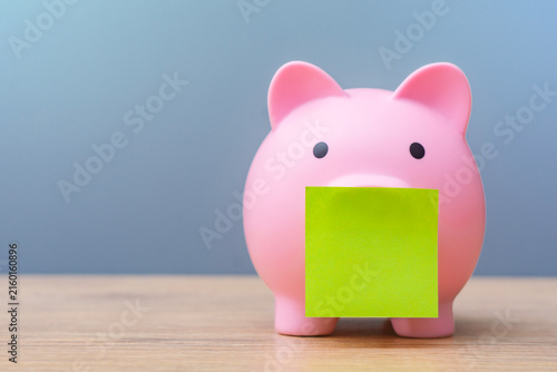 Pink piggy bank with a blank green note on a wooden table, representing savings, personal finance, budgeting, money management and financial planning with copy space.