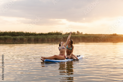 Bild auf Leinwand Teen Women Enjoying Summer Fun on a River While Paddleboarding at Sunset