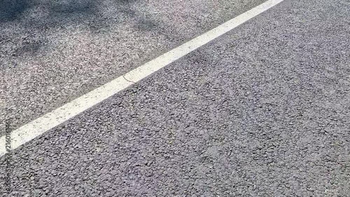 An empty gray asphalt road surface is shown close up from above, featuring a white dashed lane marking, with the camera moving steadily forward along the rough pavement texture in daylight.