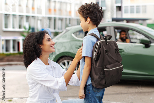 Caring mother adjusting sons backpack before school while father waiting in car