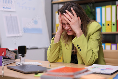 Young woman in lime blazer sits at desk covering face with hands during stressful work moment inside busy office, surrounded by laptop, papers, folders, whiteboard and binders, with stressed, stress