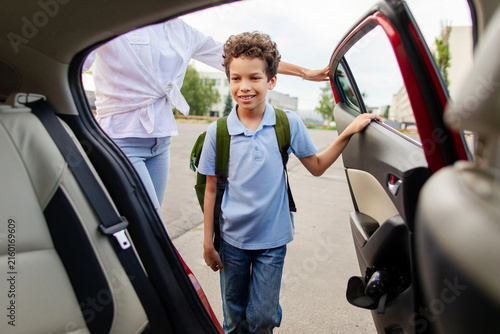 Black schoolboy entering family car after school with his mother nearby