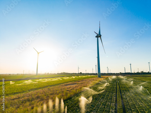 Fotografie Wind turbines stand in a green field as water sprays from irrigation systems dur