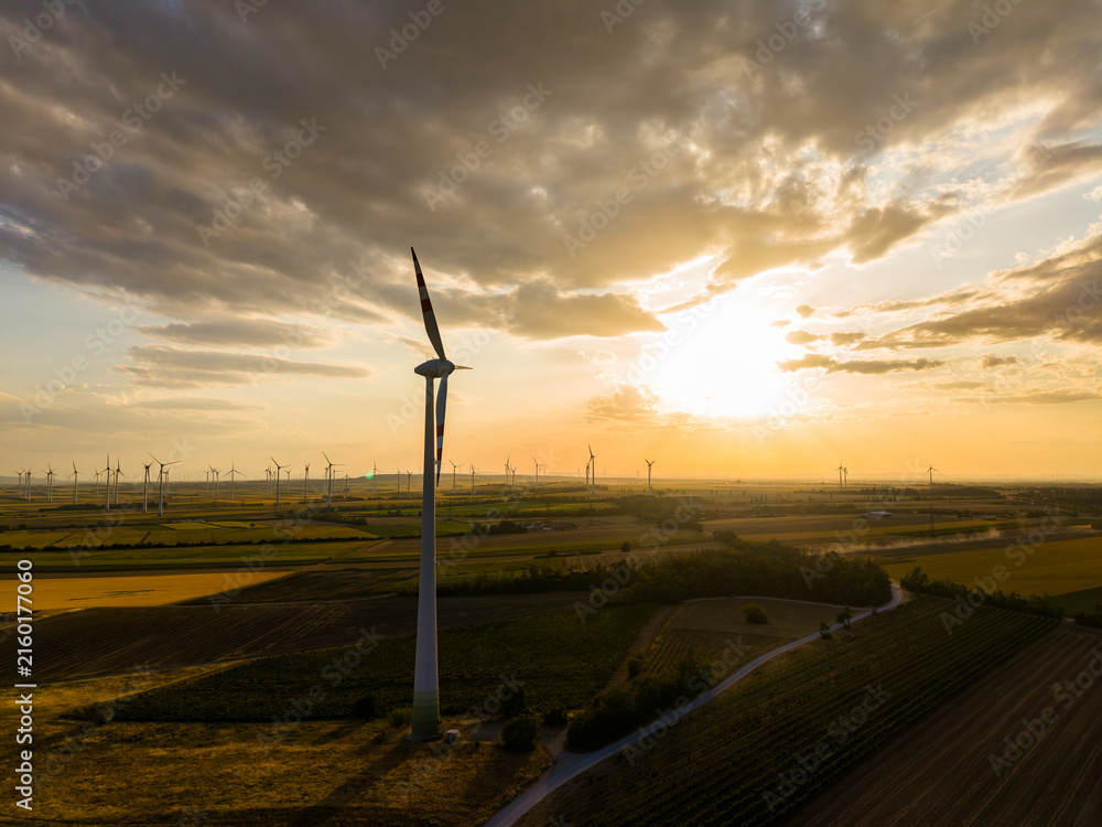 Obraz premium Wind turbines stand tall in the fields during sunset with clouds in the sky and light shining through